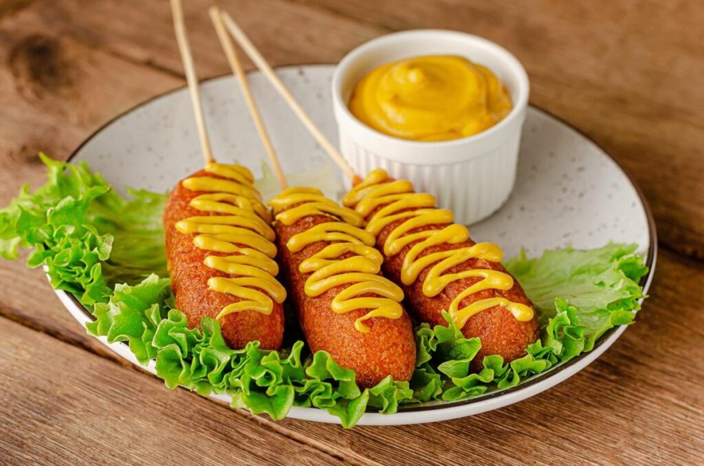 A golden Korean corn dog drizzled with chili sauce and mustard, accompanied by kimchi.
